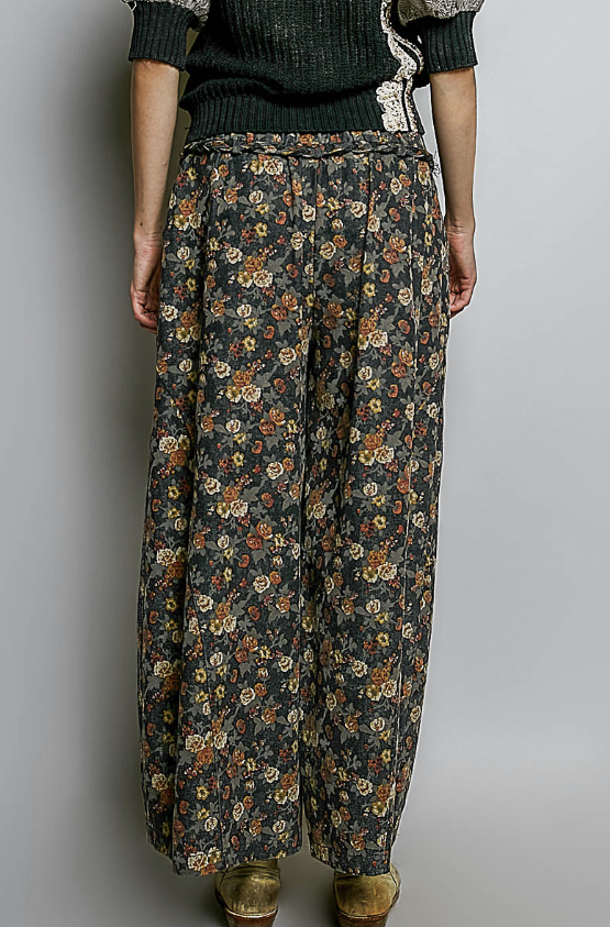 A person shown from the back wearing wide-leg floral pants in a dark gray base with orange, yellow, and cream flowers, paired with a dark green knit top and brown shoes, against a plain gray studio background.