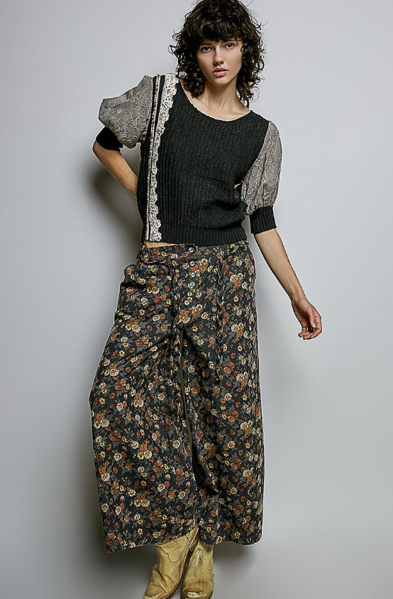 A model wearing a black ribbed knit top with sheer patterned puff sleeves and a dark floral wide-leg skirt, paired with tan boots, against a plain light gray background.