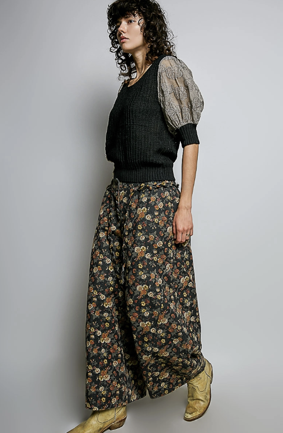 A model wearing a black ribbed knit sweater vest, sheer patterned puff sleeves, wide-leg floral pants, and tan boots, posed against a light gray studio background.