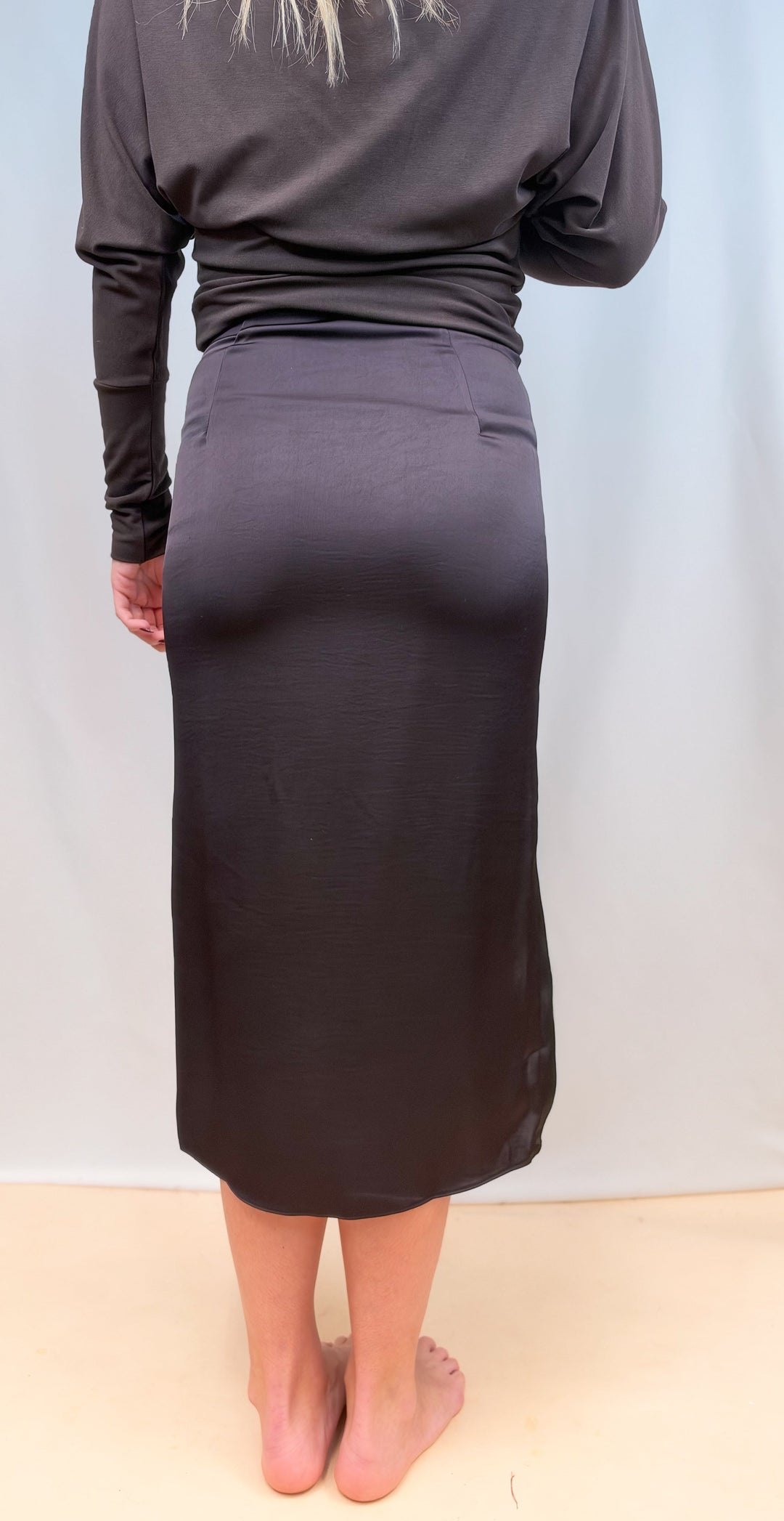 Woman shown from back view wearing brown long sleeve top and brown satin skirt on plain background