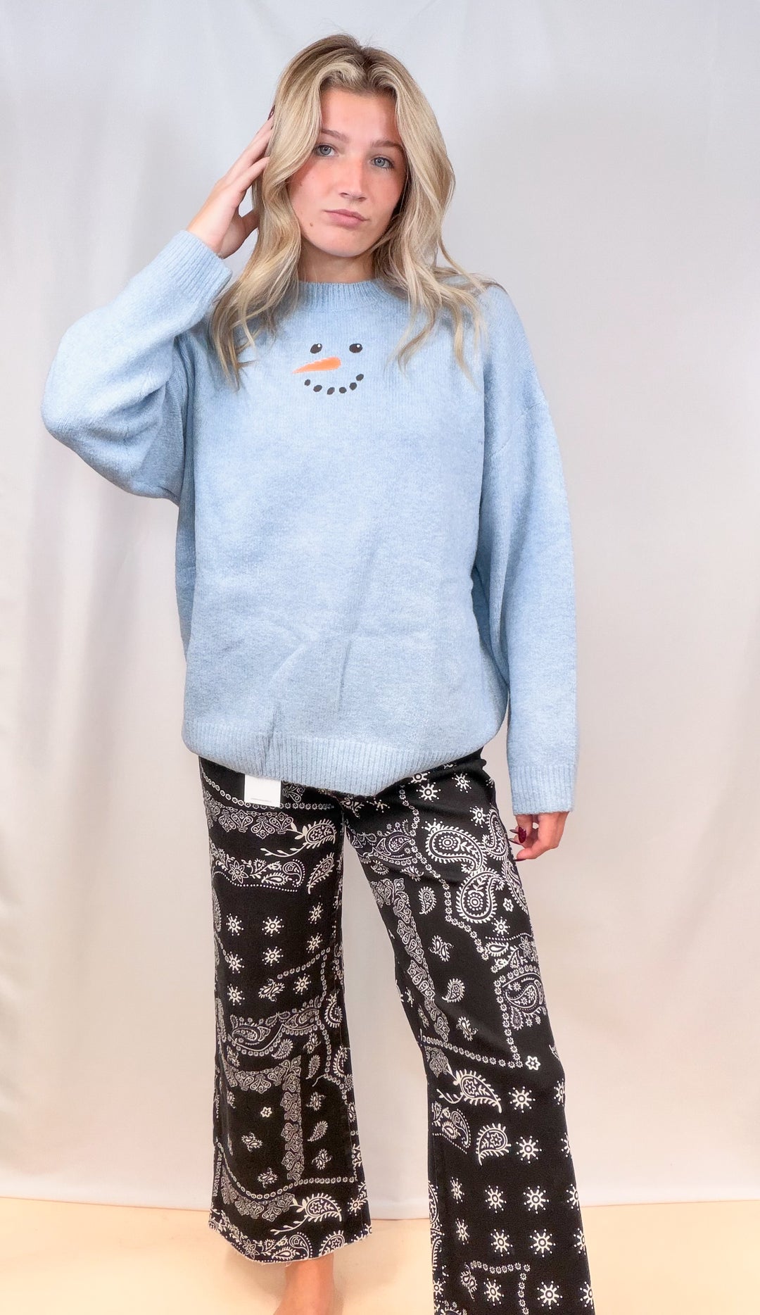 Woman wearing light blue sweater with snowman face and black bandana pants on plain background
