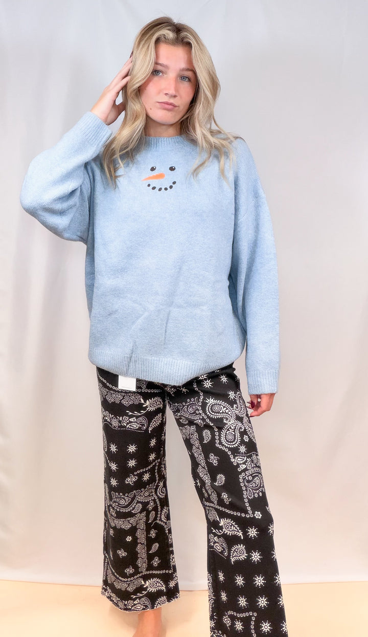 Woman wearing light blue sweater with snowman face and black bandana pants on plain background