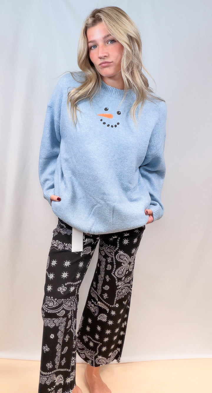 Woman wearing light blue sweater with snowman face and black bandana pants on plain background