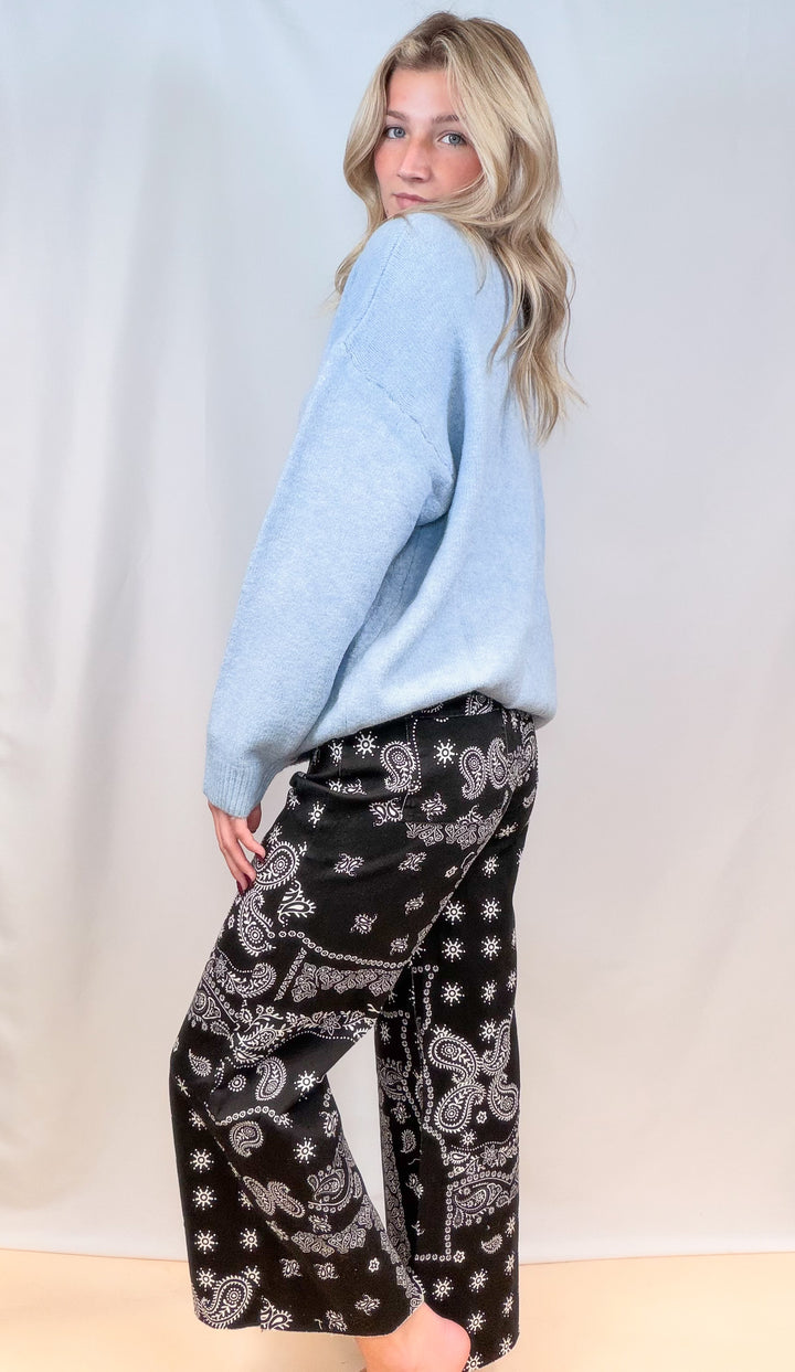 Woman shown from side view wearing light blue sweater and black bandana pants on plain background