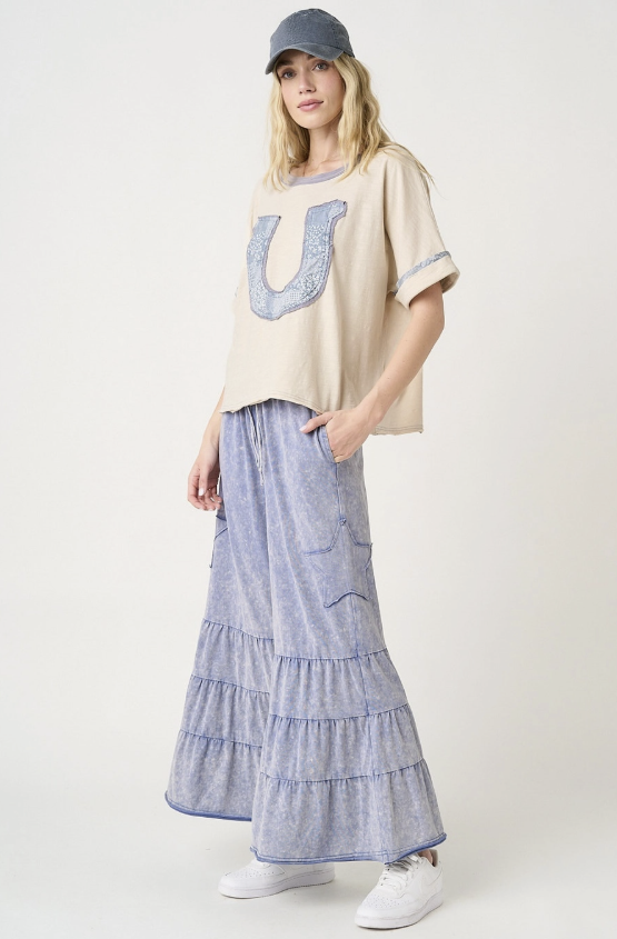 Woman wearing a beige oversized shirt with a blue 'U' design, a blue tiered skirt, and white sneakers on a white background.