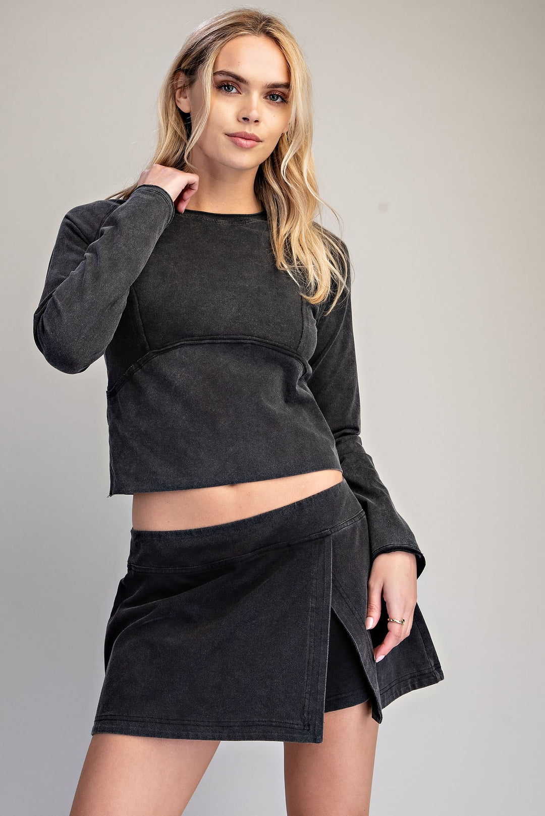 A blonde model wearing a dark charcoal gray cropped long-sleeve top and a matching dark gray wrap-style mini skirt, photographed against a neutral light gray studio background.