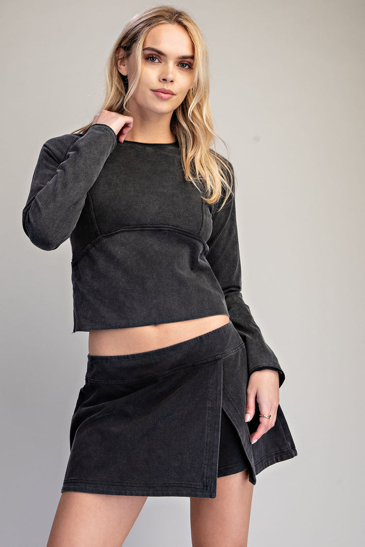 A blonde model wearing a dark charcoal gray cropped long-sleeve top and a matching dark gray wrap-style mini skirt, photographed against a neutral light gray studio background.