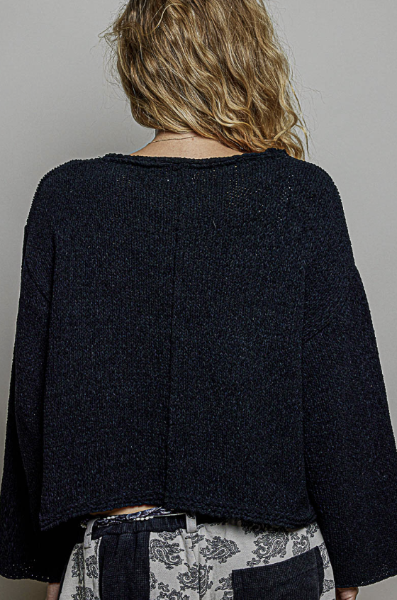 Back view of a person wearing a dark navy blue cropped knit sweater and patterned gray pants against a plain gray background.