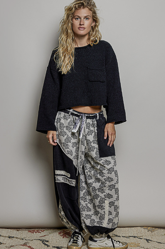 A woman modeling a black cropped knit long-sleeve top and patterned black-and-white wide-leg pants with a tie belt, standing against a plain light gray wall.