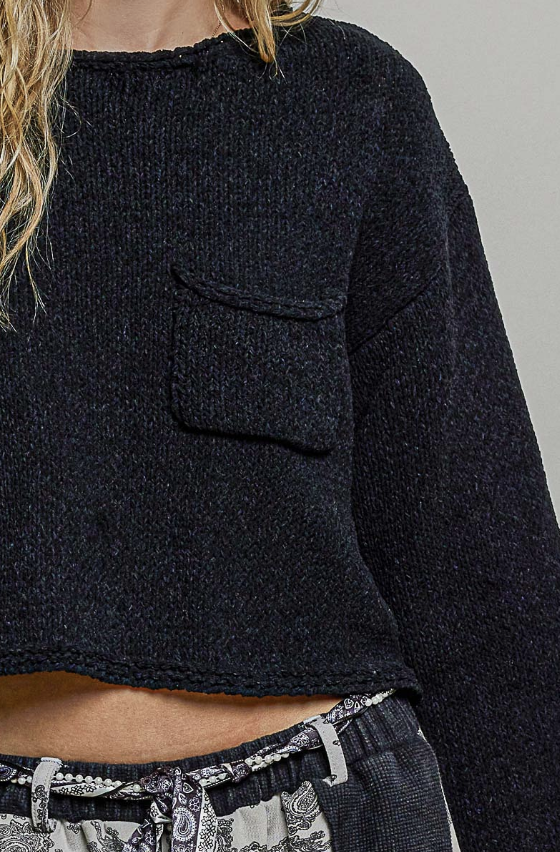 Close-up of a person wearing a dark charcoal knit cropped sweater with a chest pocket, paired with patterned gray and white bottoms.