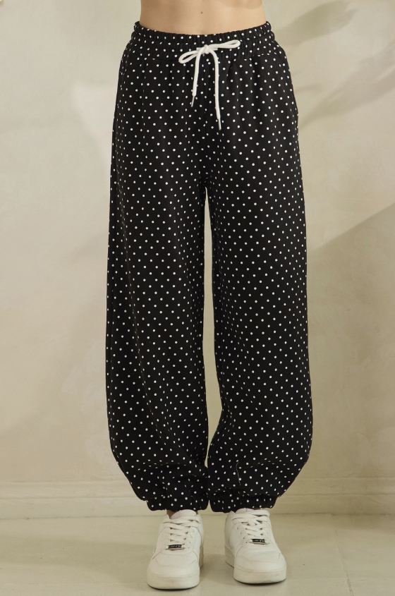 Black wide-leg pants with a white polka dot pattern and a white drawstring waist, shown on a model wearing white sneakers against a neutral beige background.