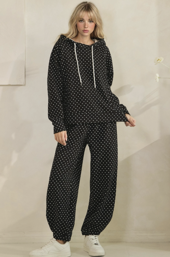 A woman modeling a matching black and white polka dot loungewear set, featuring a long-sleeve hooded sweatshirt with white drawstrings and matching wide-leg jogger pants, styled with white sneakers.