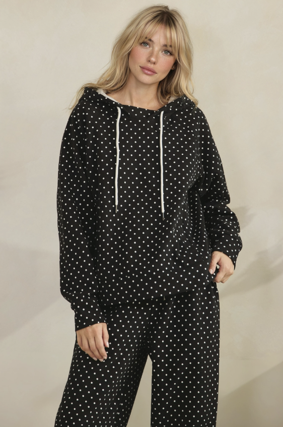 A woman modeling a matching black and white polka dot lounge set, featuring a long-sleeve hooded sweatshirt with white drawstrings and matching wide-leg pants, set against a plain beige studio background.