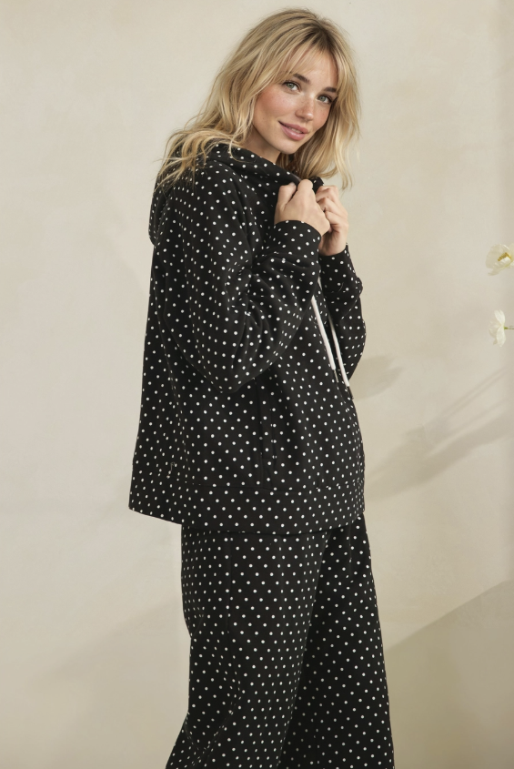 A woman modeling a matching black and white polka dot lounge set, featuring a long-sleeve top and wide-leg pants, against a plain beige wall.