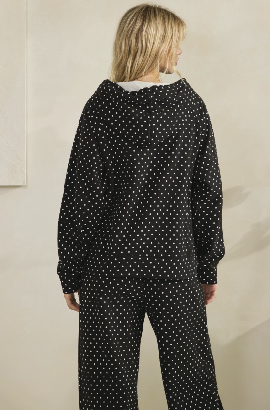 A model viewed from the back wearing a black long-sleeve jumpsuit with a white polka dot pattern and a white collar, set against a plain beige wall.