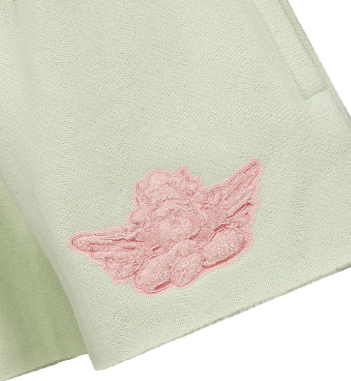 Close-up of a pale sage green fabric item featuring a pink embroidered cherub motif on the lower left side.