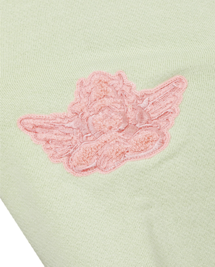 A close-up of a pale sage green knit fabric featuring a pink embroidered angel wing applique.