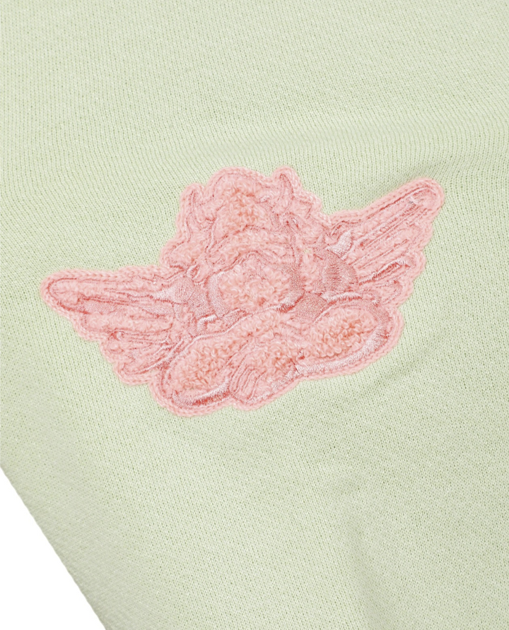 A close-up of a pale sage green knit fabric featuring a pink embroidered angel wing applique.