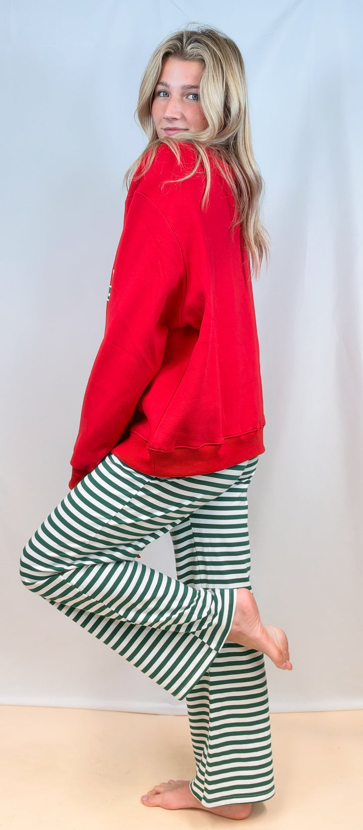 Woman shown from side view wearing red sweatshirt with green and white stripe pants on plain background