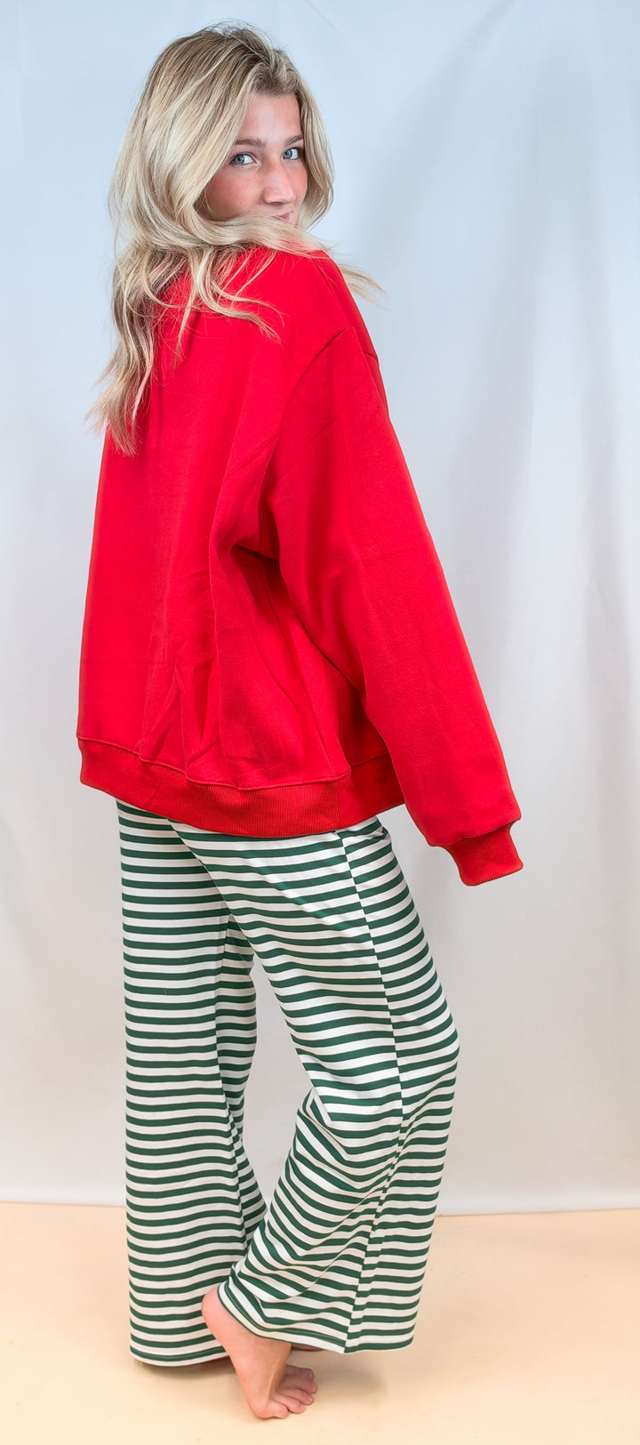 Woman shown from back wearing red sweatshirt with green and white stripe pants on plain background