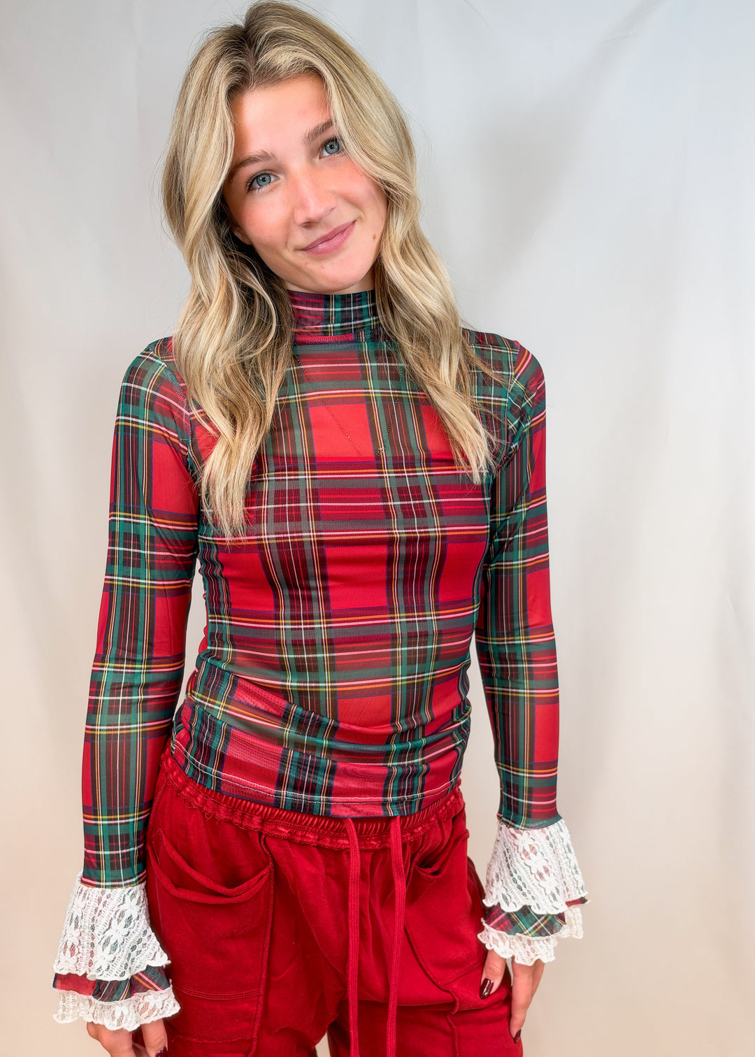 Woman wearing red and green tartan sheer shirt with lace detail cuffs and red pants on plain background