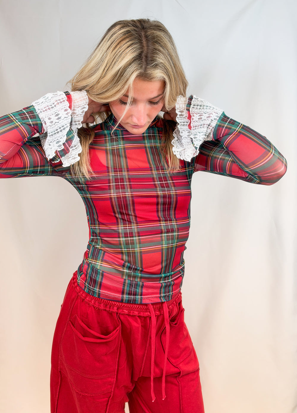 Woman wearing red and green tartan sheer shirt with lace cuff detail and red pants on plain background