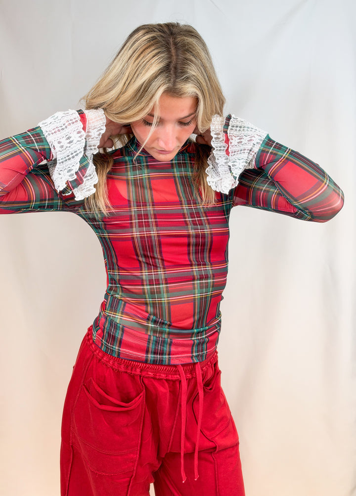 Woman wearing red and green tartan sheer shirt with lace cuff detail and red pants on plain background