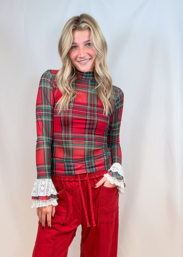 Woman wearing red and green tartan sheer shirt with lace cuff details and red pants on plain background
