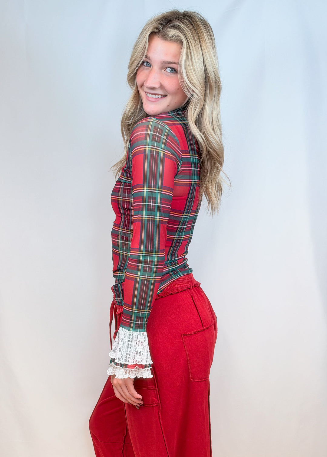 Woman from side view wearing red and green tartan sheer shirt with lace cuff detail and red pants on plain background