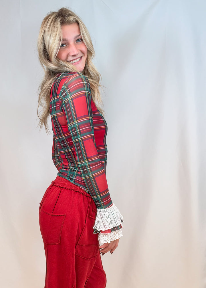 Woman shown from side view wearing red and green tartan sheer shirt with lace cuff sleeves and red pants on plain background