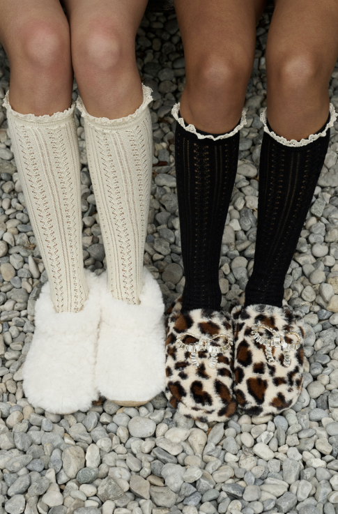 Two pairs of knee-high socks and slippers shown on legs standing on gray gravel. The left pair consists of cream cable-knit socks with white fluffy slippers.