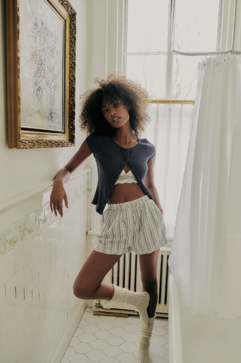 A model with curly hair stands in a bathroom setting, wearing a dark gray cropped top, striped shorts, and white socks.