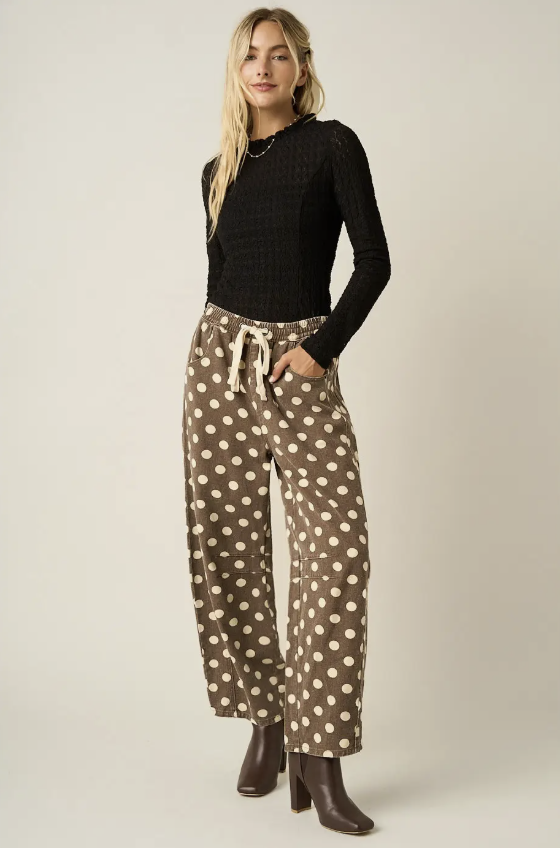A woman modeling a black textured long-sleeve top and brown wide-leg pants with beige polka dots, paired with dark brown ankle boots, against a solid light beige studio background.