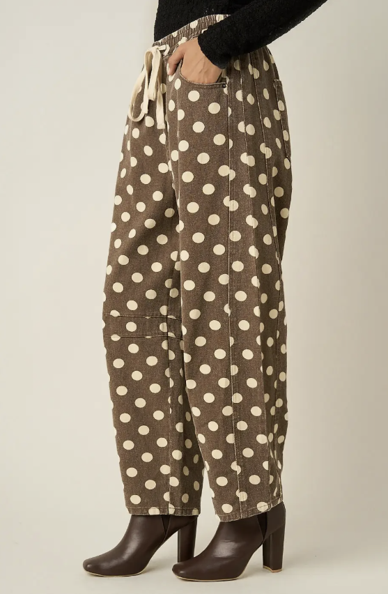 A person wearing brown wide-leg pants with a cream polka dot pattern, a cream drawstring waist, and dark brown heeled ankle boots, shown from the waist down against a light beige studio background.