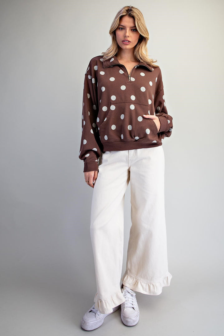 A woman modeling a brown long-sleeve half-zip sweatshirt with white polka dots, paired with off-white wide-leg ruffled pants and white sneakers, against a neutral gray studio background.