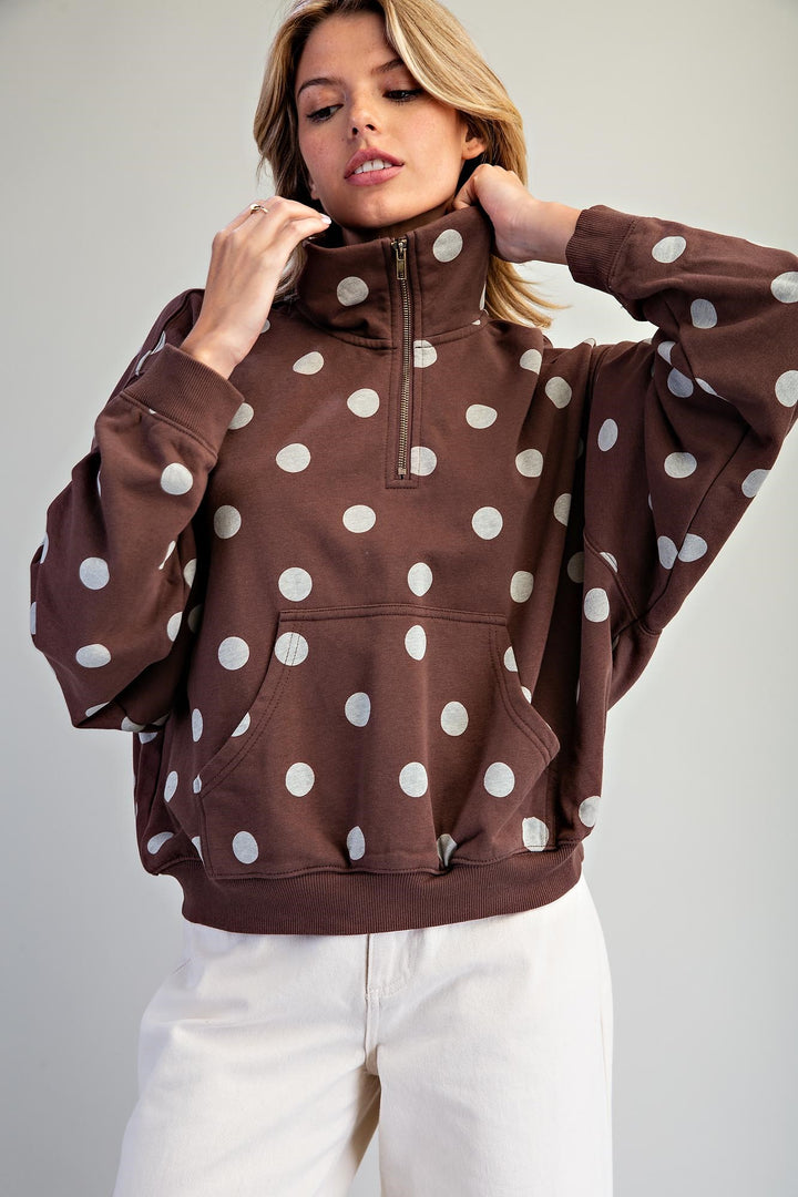 A woman wearing a brown half-zip pullover sweatshirt with a white polka dot pattern, paired with white pants, against a plain light gray background.