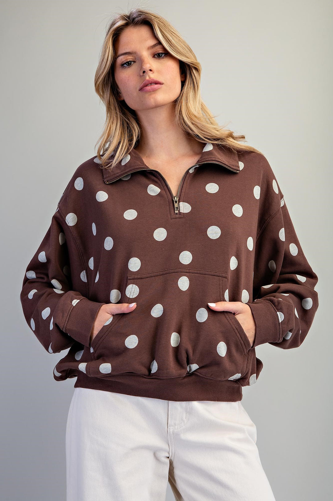 A woman wearing a brown half-zip sweatshirt with white polka dots and white pants, standing against a light gray studio background.