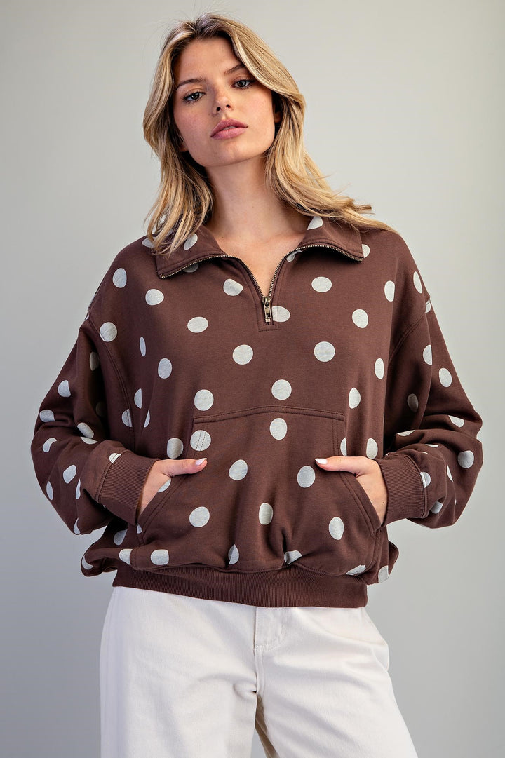 A woman wearing a brown half-zip sweatshirt with white polka dots and white pants, standing against a light gray studio background.