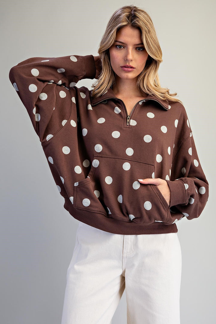 A woman wearing a brown half-zip sweatshirt with large white polka dots and white pants, posing against a neutral light gray background.