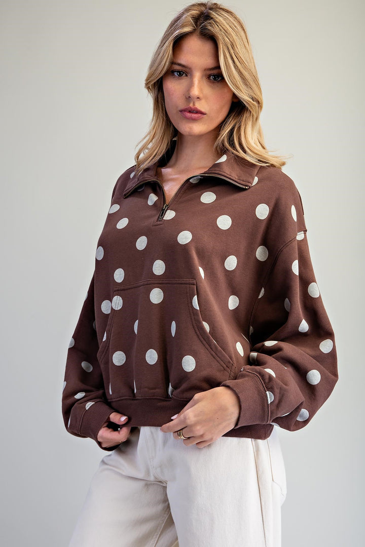 A woman wearing a brown half-zip sweatshirt with large white polka dots, paired with off-white pants, photographed against a neutral light gray studio background.