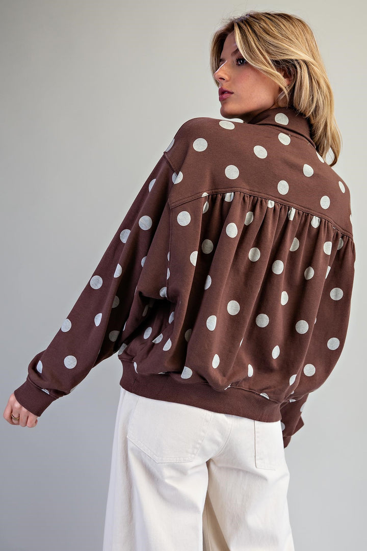 A woman wearing a brown long-sleeve top with large white polka dots and gathered ruffle details, paired with white pants, photographed against a plain light gray studio background.