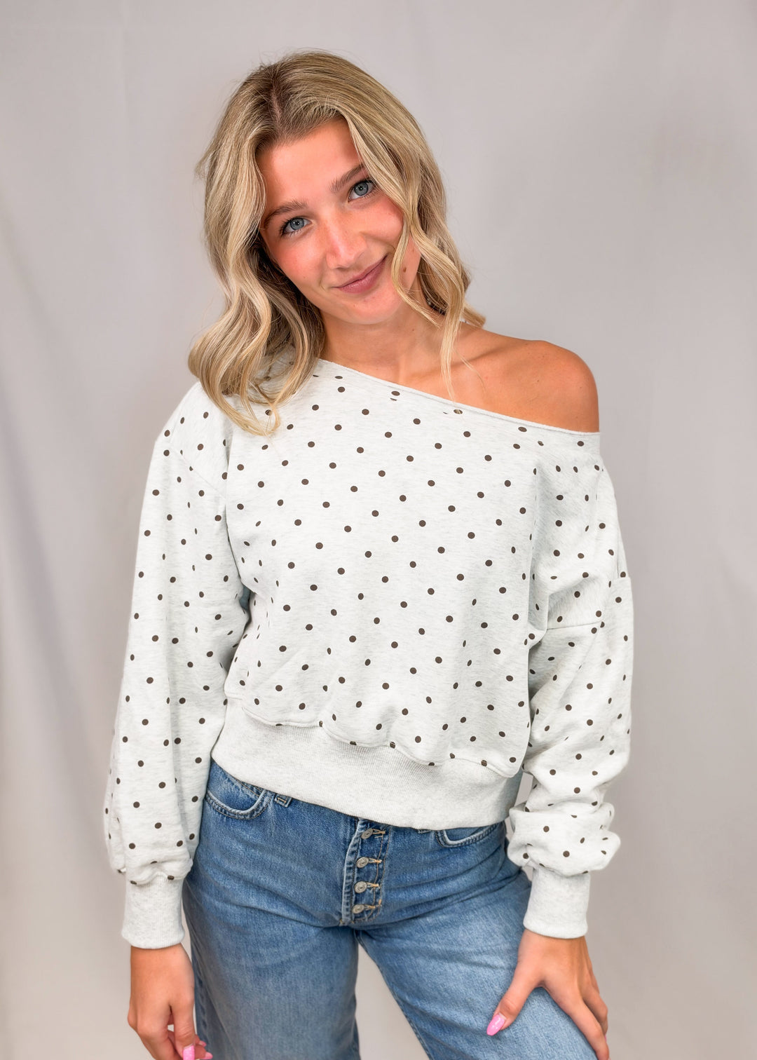 Woman wearing heather grey off shoulder sweatshirt with brown polka dots and jeans on plain background