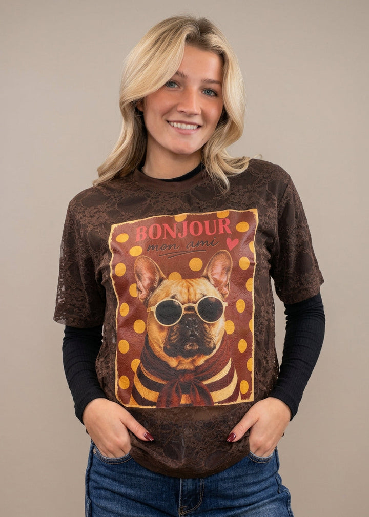 Woman wearing brown lace tee with black long sleeve under. Tee has french bulldog graphic on front with denim jeans on plain background
