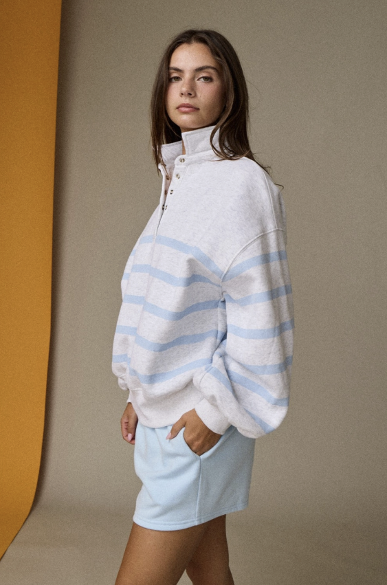 A woman modeling a white half-zip sweatshirt with light blue horizontal stripes, paired with a light blue mini skirt, set against a neutral studio backdrop with a yellow-orange color block on the left.