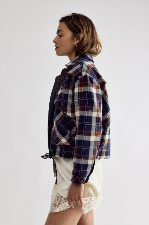 A woman shown in profile wearing a cropped plaid jacket over a dark top and cream lace shorts, photographed against a plain light gray studio background.