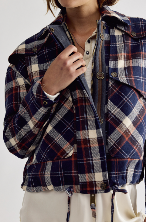 A close-up of a person wearing a cropped plaid jacket in navy, cream, and rust red tones, layered over a cream shirt. The jacket features a zippered front, snap buttons, and a plaid pattern.