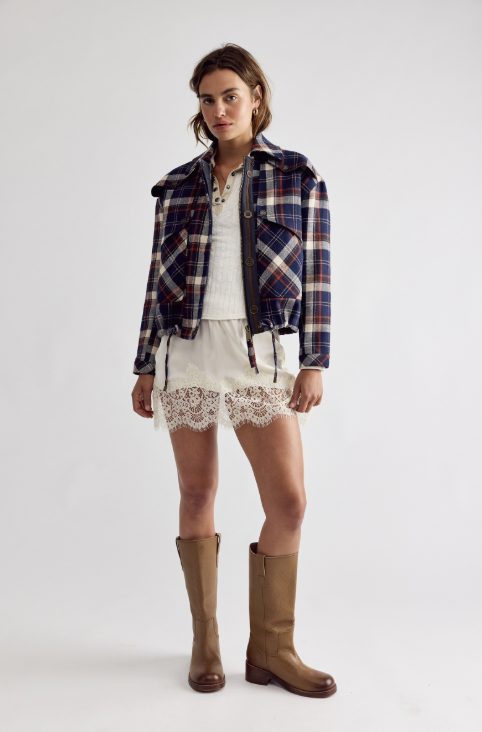 A model stands against a light gray studio background wearing a navy and red plaid jacket over a white ribbed top and white lace-trim shorts, paired with brown knee-high boots.