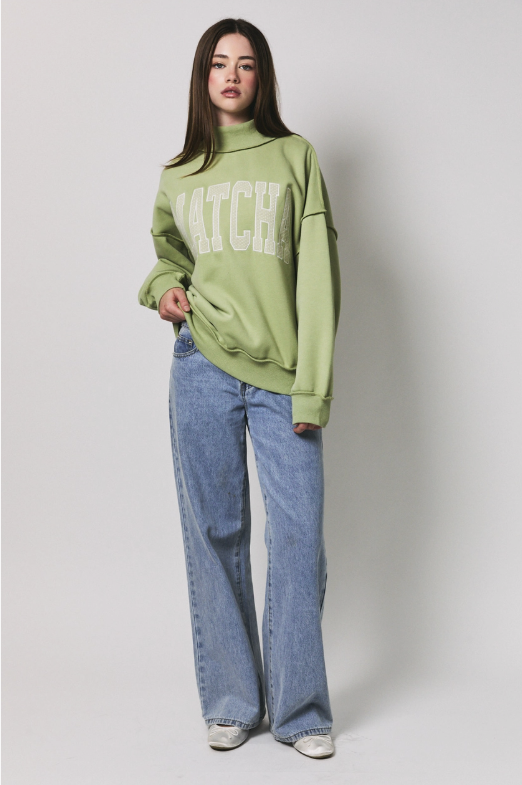 A woman modeling a sage green mock-neck sweatshirt with 'MATCHA' printed in white across the chest, paired with light-wash wide-leg jeans and white sneakers, against a plain light gray studio background.