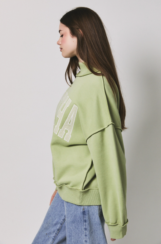 A woman in profile wearing a light sage green sweatshirt with large white textured lettering on the back, paired with light blue jeans, against a plain light gray studio background.