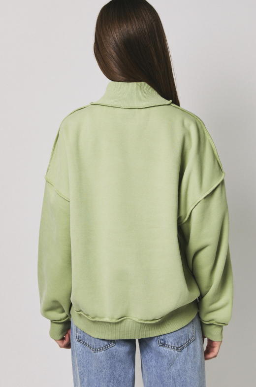 A woman viewed from the back wearing an oversized sage green mock-neck sweatshirt and light blue jeans against a plain light gray studio background.